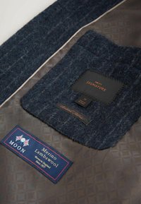 Navyblauw wollen stof met een gestructureerde afwerking, met labels: één zwart met "NEXT SIGNATURE" en een andere blauwe voor "Moon Merino Lambswool."