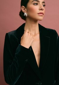 Must must-blazer sügava dekoltee, ühe nupuga sulgemine ja üleskeeratud varrukad. Mudel kannab tekstuuriga hõbedast kõrvarõngast roosa tausta ees.