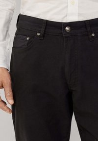 Zwarte katoenen jeans met een slanke pasvorm, voorzien van standaard zakken en metalen details, waaronder een knoop en klinknagels. Glad oppervlak.