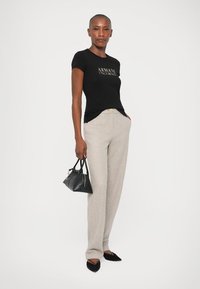 Tricou negru mulat cu logo metalic, pantaloni largi bej cu model, geantă neagră și pantofi negri cu vârf, evidențiind un stil minimalist.