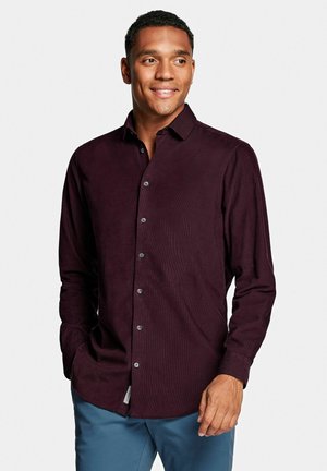 Man die lacht en naar rechts kijkt, draagt een donkerpaars overhemd met lange mouwen en blauwe broek, staand tegen een effen achtergrond.