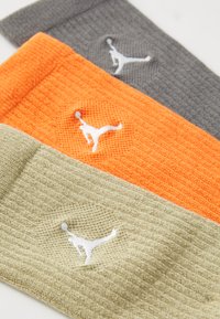 Tres cintas deportivas: naranja, gris y verde oliva, con un pequeño logo blanco, tela texturizada y diseño acanalado.