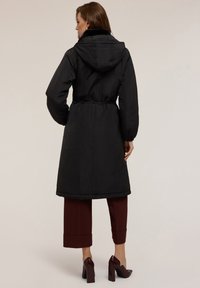 Svart parkas med huva, insvängd midja och elastiska ärmslut. Har en mjuk krage och knälång modell, bärs över vinröda korta byxor.