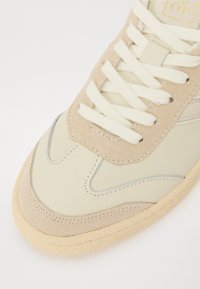 Beige sneaker med en glat læderoverdel og suedeblæk, med hvide snørebånd, en tekstureret sål og subtile stikninger.