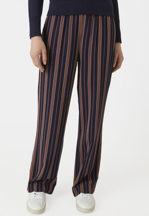 Persoon die marineblauwe, bruine en beige verticale gestreepte wijde broek draagt met witte sneakers en een donker shirt met lange mouwen.