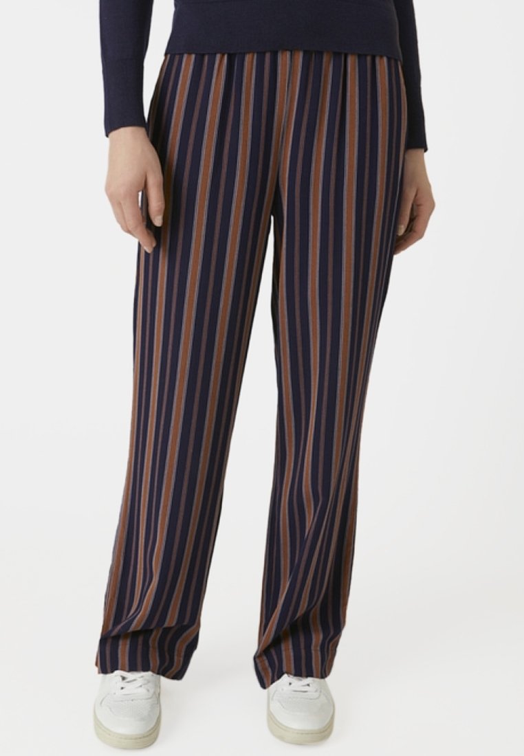 Personne portant un pantalon à jambes larges à rayures verticales bleu marine, marron et beige, avec des baskets blanches et un haut sombre à manches longues.