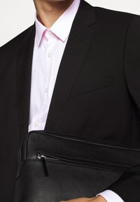 Czarna skórzana torba na laptopa z zamkiem błyskawicznym, trzymana na tle czarnego garnituru i jasnoróżowej koszuli.