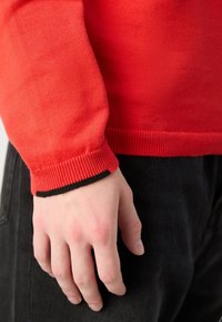 Gros plan d’une main gauche à côté de la hanche portant un pull rouge avec un poignet bordé de noir et un pantalon noir.