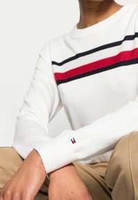 Maglione bianco a maniche lunghe con strisce orizzontali rosse e blu navy sul petto. I polsini a coste presentano un piccolo logo decorativo.