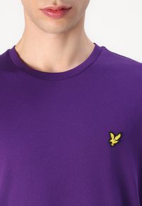 Violets krekls no kokvilnas ar apaļu kakla izgriezumu. Ar dzeltenu izšūtu logo kreisajā krūtīs. Gluda tekstūra.
