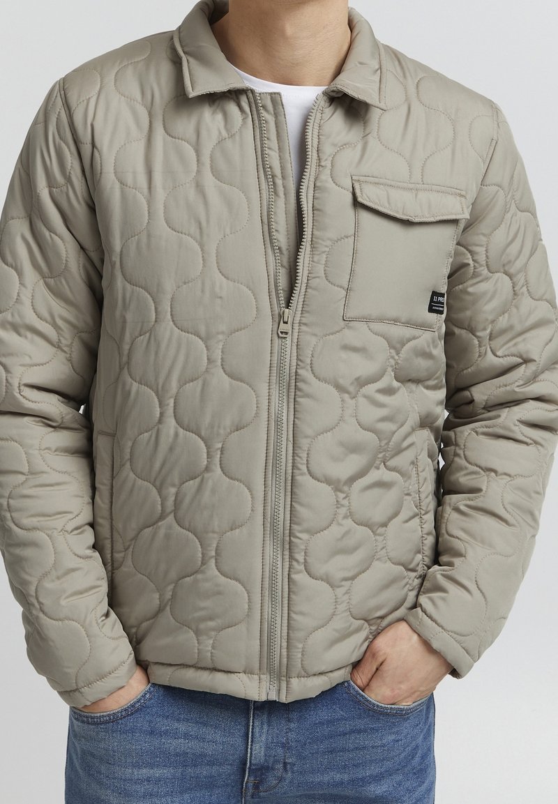 Veste matelassée beige avec un motif texturé en ondulations, dotée d'une fermeture éclair sur le devant et d'une poche poitrine. Conçue avec un col classique.