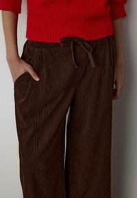 Brązowe szerokie spodnie z sztruksu z bocznymi kieszeniami i brązowym sznurkiem w pasie. Tkanina ma pionową, żebrowaną strukturę.