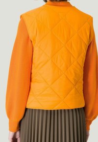 Gilet matelassé orange avec un motif en losanges, associé à un pull orange à côtes. Jupe plissée vert foncé aperçue en bas.