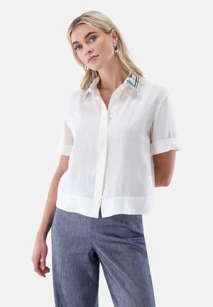 Blonde woman wearing white short-sleeve button-up shirt with striped collar and blue high-waisted pants, standing against plain background.