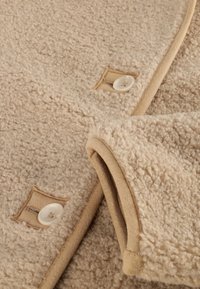 Beige fleece stof met een zachte, gestructureerde afwerking, voorzien van een tan rand en knopenaccenten nabij de gestikte zakken.