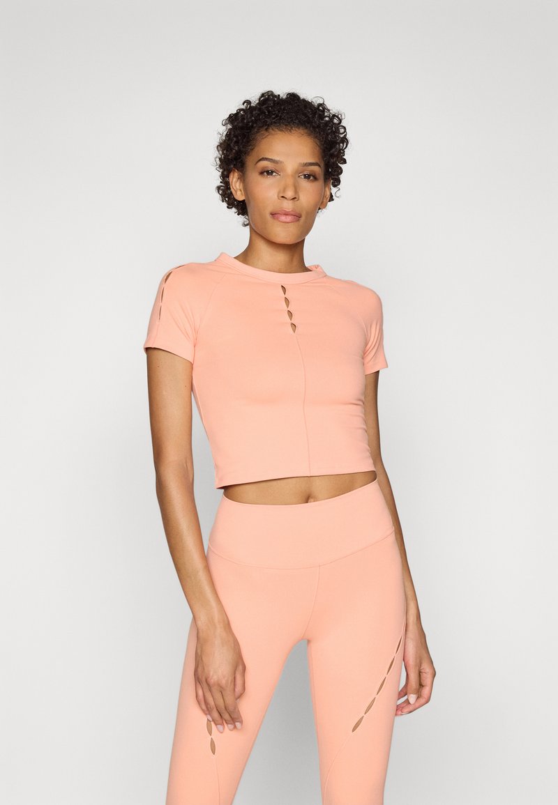 Pfirsichfarbener Crop-Top mit kurzen Ärmeln, hochgeschlossen und mit Ausschneidungen; kombiniert mit passenden, hochgeschnittenen Leggings mit seitlichen Schlitzen.