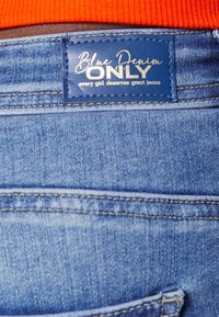 Jeans de ganga azul com uma etiqueta de couro azul escura rotulada "ONLY" em escrita branca, realçada com costura e uma textura suave.