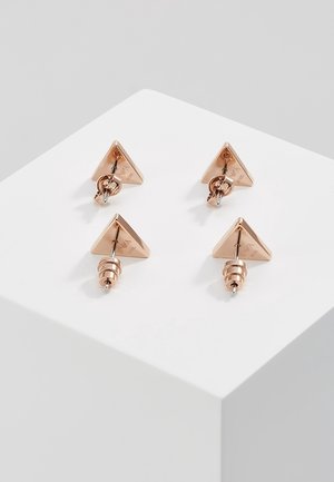 Quatre boucles d’oreilles clous triangulaires en or rose avec fermoirs en spirale exposées sur un cube blanc sur fond gris clair.