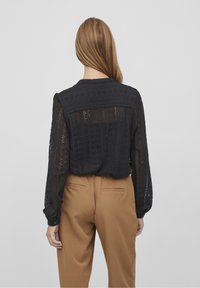 Blouse en dentelle noire présentant un design texturé, des manches longues et un encolure arrondie. Associée à un pantalon beige et à une silhouette droite.