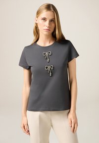 Camiseta gris de algodón, con dos lazos decorativos con acentos brillantes en el centro. Manga corta y corte relajado.