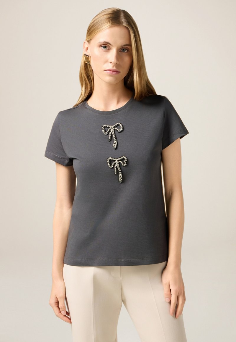 Camiseta gris de algodón, con dos lazos decorativos con acentos brillantes en el centro. Manga corta y corte relajado.