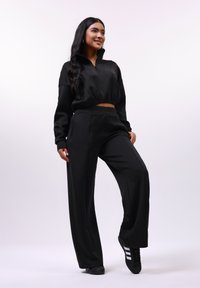 Jonge vrouw die glimlacht, gekleed in een zwart zip-up crop top, wijdvallende broek en zwarte sneakers met witte strepen, staand met één been iets opgetild.