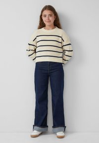 Crème gebreide trui met marineblauwe strepen, gecombineerd met flare donkerblauwe jeans en witte sneakers, staand tegen een neutrale achtergrond.