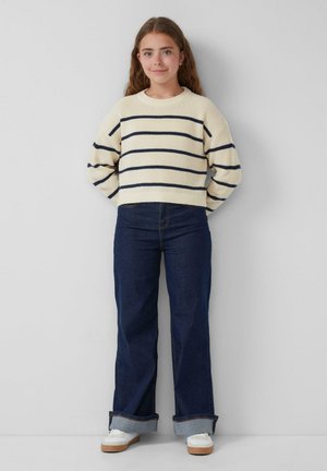 Cremfarbener Strickpullover mit marineblauen Streifen, kombiniert mit ausgestellten dunkelblauen Jeans und weißen Sneakers, vor einem neutralen Hintergrund stehend.