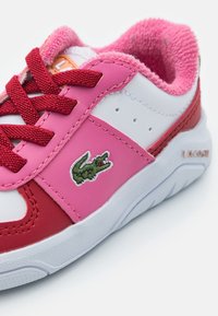 Tenisky Lacoste s bílým a růžovým syntetickým svrškem, červenými detaily, texturovaným růžovým límcem a vyšitým krokodýlem.