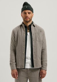 Camicia a quadri in tonalità beige e marrone, con taschino, indossata sopra una t-shirt bianca, abbinata a pantaloni coordinati e un berretto verde.