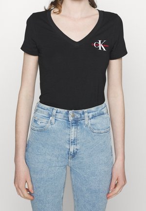 Black v-neck t-shirt made of cotton, featuring a red and white logo on the left chest, paired with light blue high-waisted jeans.