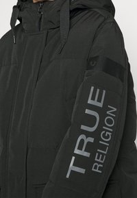 Svart puffjacka med huva, dragkedja och sidofickor. En framträdande "TRUE RELIGION"-logotyp i grått på ärmen. Slät textur.