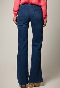 Blå utsvängda jeans i denim, med hög midja, två bakfickor och en lätt lyster. Tyget har en slät och mjuk textur.