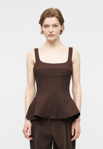 Femme aux cheveux châtain clair courts portant un haut péplum marron sans manches ajusté et un pantalon assorti, debout devant un fond uni.