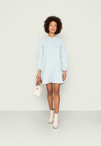 Vestido listrado em azul claro, com bainha franzida e mangas longas, apresentando um decote redondo e detalhes em botão. A modelo segura uma bolsa branca.