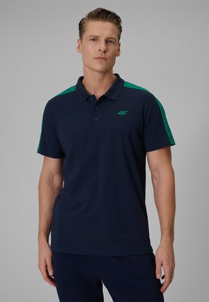 Mężczyzna noszący granatową koszulkę polo z zielonymi paskami na ramionach i małym logo na lewej piersi na szarym tle.