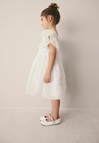 Weißes gestuftes Kleid mit Rüschenärmeln, aus leichtem Stoff. Kind trägt weiße Mary-Jane-Schuhe mit Schnallendetail, Seitenansicht.