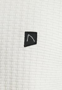 Weißer strukturierter Stoff mit eingewebtem Muster, mit einem kleinen schwarzen Etikett, das ein weiß umrandetes Design enthält und im Stoff eingearbeitet ist.
