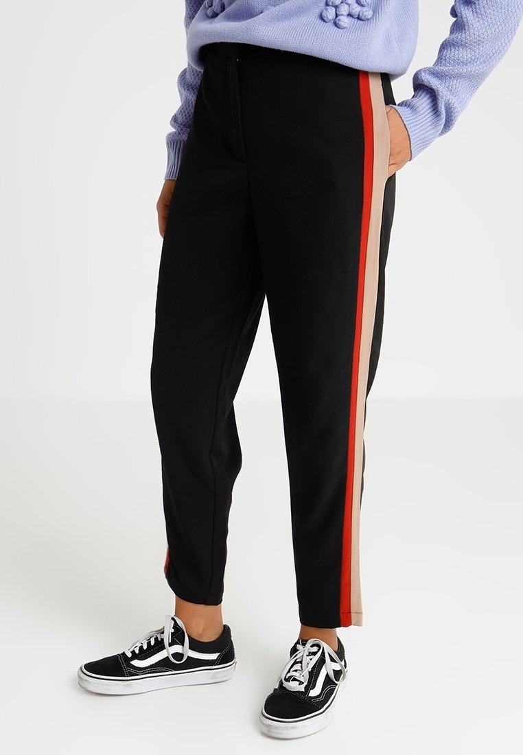 Osoba nosząca czarne tapered pants z czerwonymi i beżowymi paskami po bokach, w połączeniu z czarno-białymi sneakersami oraz jasnofioletowym swetrem