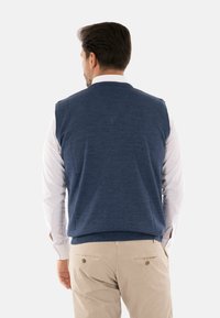 Maglione senza maniche blu navy in maglia lavorata, indossato sopra una camicia bianca a maniche lunghe, abbinato a pantaloni chiari. Vista dal retro.