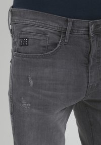 Jeans de mezclilla gris con acentos desgastados, que cuentan con un bolsillo frontal con una etiqueta negra y un diseño clásico de cinco bolsillos.