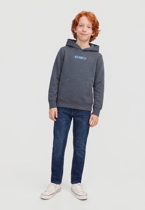 Rothaariger Junge mit dunklegrauem Hoodie, blauen Jeans und weißen Sneakers, der mit einem leichten Lächeln vor einem einfarbigen hellen Hintergrund steht.