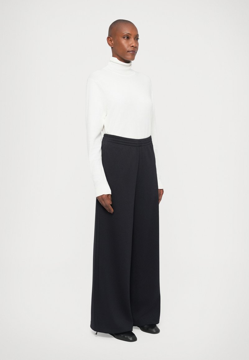 Personne avec la tête rasée, portant un col roulé blanc et un pantalon noir à large jambe, se tenant devant un fond clair et uni.