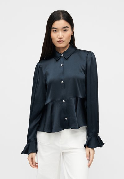 Γυναίκα με μακριά σκούρα μαλλιά που φοράει σκούρο ναυτικό μπλε πουκάμισο με κουμπιά και φαρδιές μανσέτες, μαζί με άσπρο παντελόνι με ψηλή μέση, σε ουδέτερο φόντο.