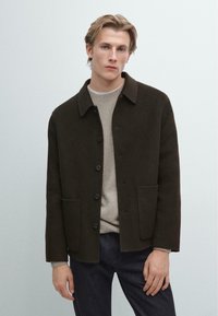 Giacca in misto lana marrone con colletto, bottoni frontali e due tasche. Indossata sopra un maglione di cotone leggero e abbinata a jeans scuri.