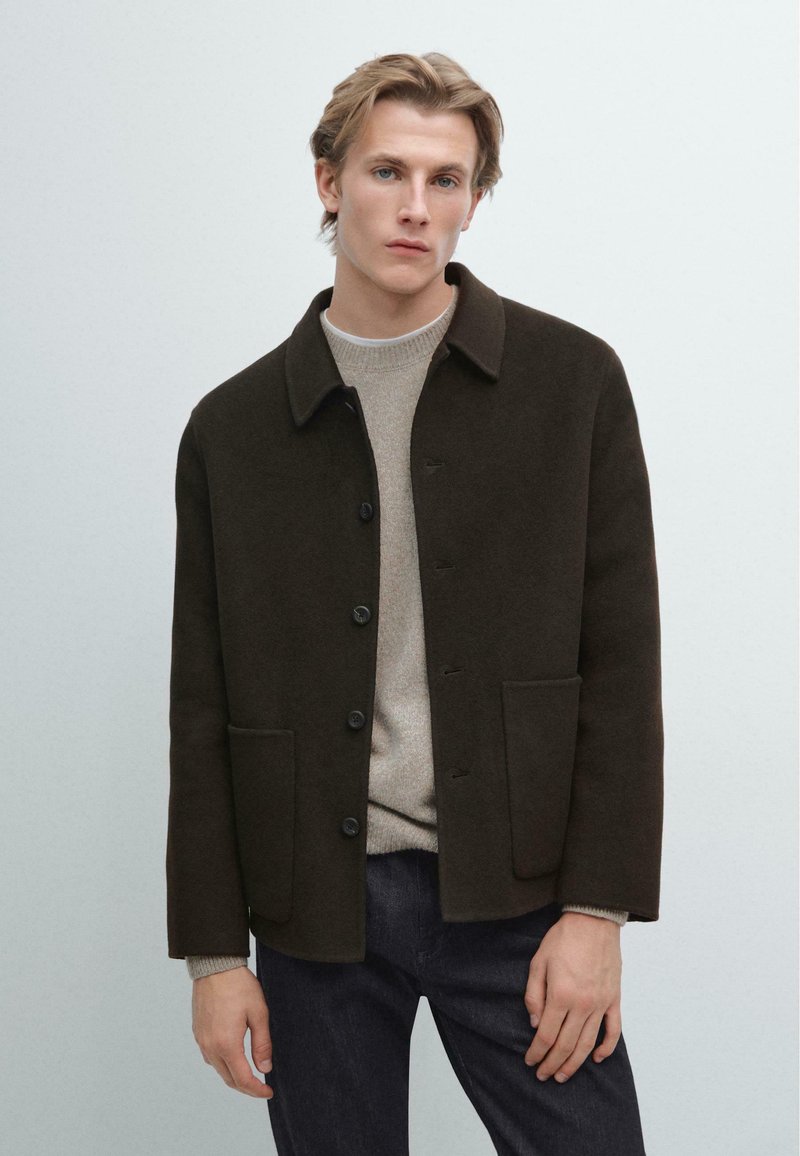 Giacca in misto lana marrone con colletto, bottoni frontali e due tasche. Indossata sopra un maglione di cotone leggero e abbinata a jeans scuri.
