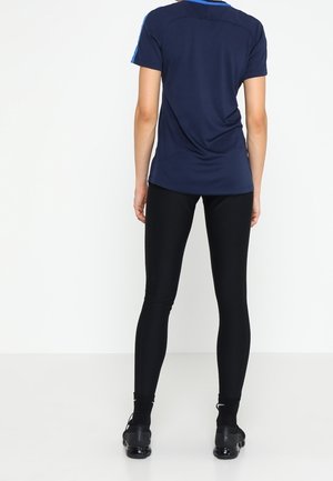 Personne debout, de dos, portant un t-shirt bleu marine à manches courtes, des leggings noirs et des chaussures de sport noires sur un fond blanc.