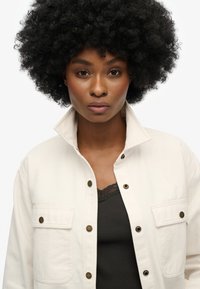 Chaqueta de lona beige con botones a presión, dos bolsillos en el pecho, llevada sobre una camiseta sin mangas con encaje negro. La modelo tiene un peinado rizado y voluminoso.