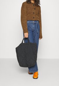 Svart vadderad tote-väska med rundade mönster, korta handtag och en mjuk textur; bärs med en brun tröja och blå jeans.
