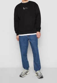 Felpa nera con logo bianco sul petto, abbinata a jeans dritti blu medio e sneakers grigie, in piedi di fronte a uno sfondo neutro.